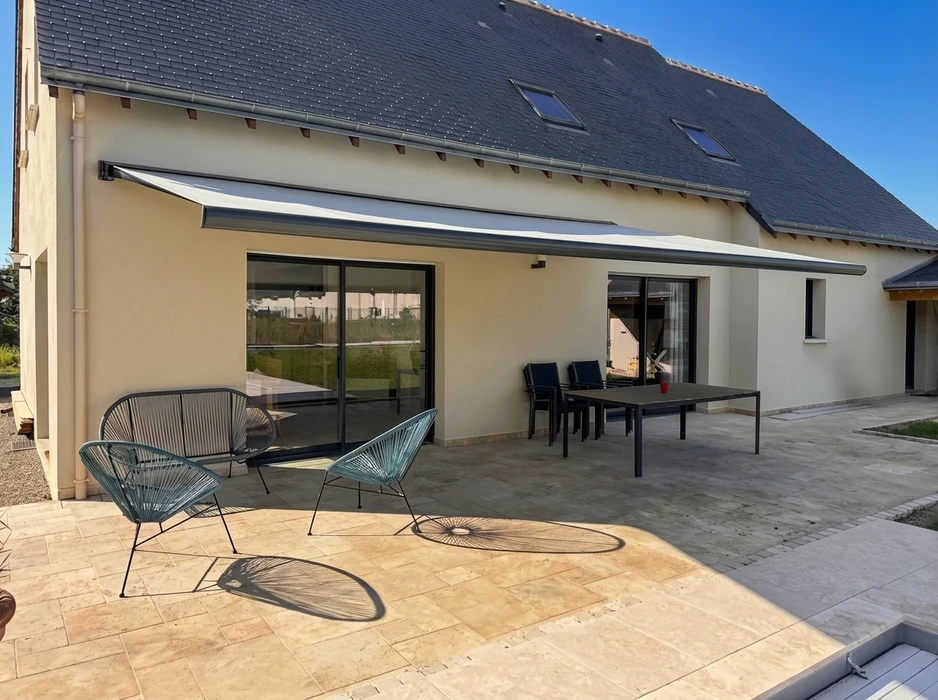 Terrasse extérieure pavée en travertin, équipée d'un store banne motorisé à coffre intégral, toile bicolore blanc/gris anthracite, fixé en façade enduite