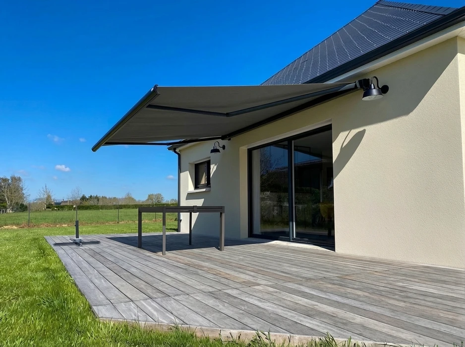 Store banne rétractable gris anthracite installée en façade d'une maison contemporaine enduit beige, au-dessus d'une terrasse en bois composite